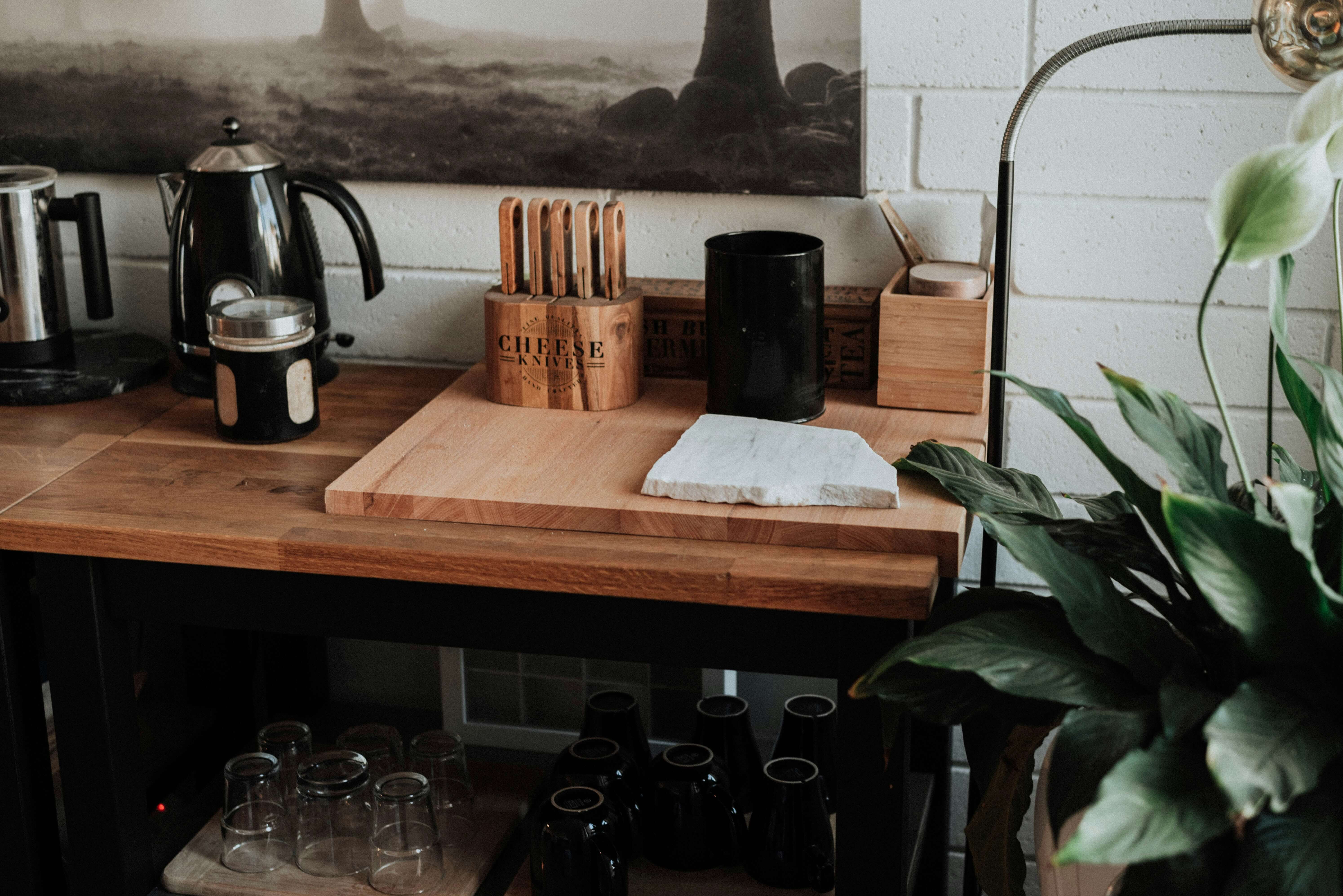 Cozy in-home coffee bar with coffee pots, cups, utensils, and a large lily plant, set against a white brick wall with a landscape photo hanging above.